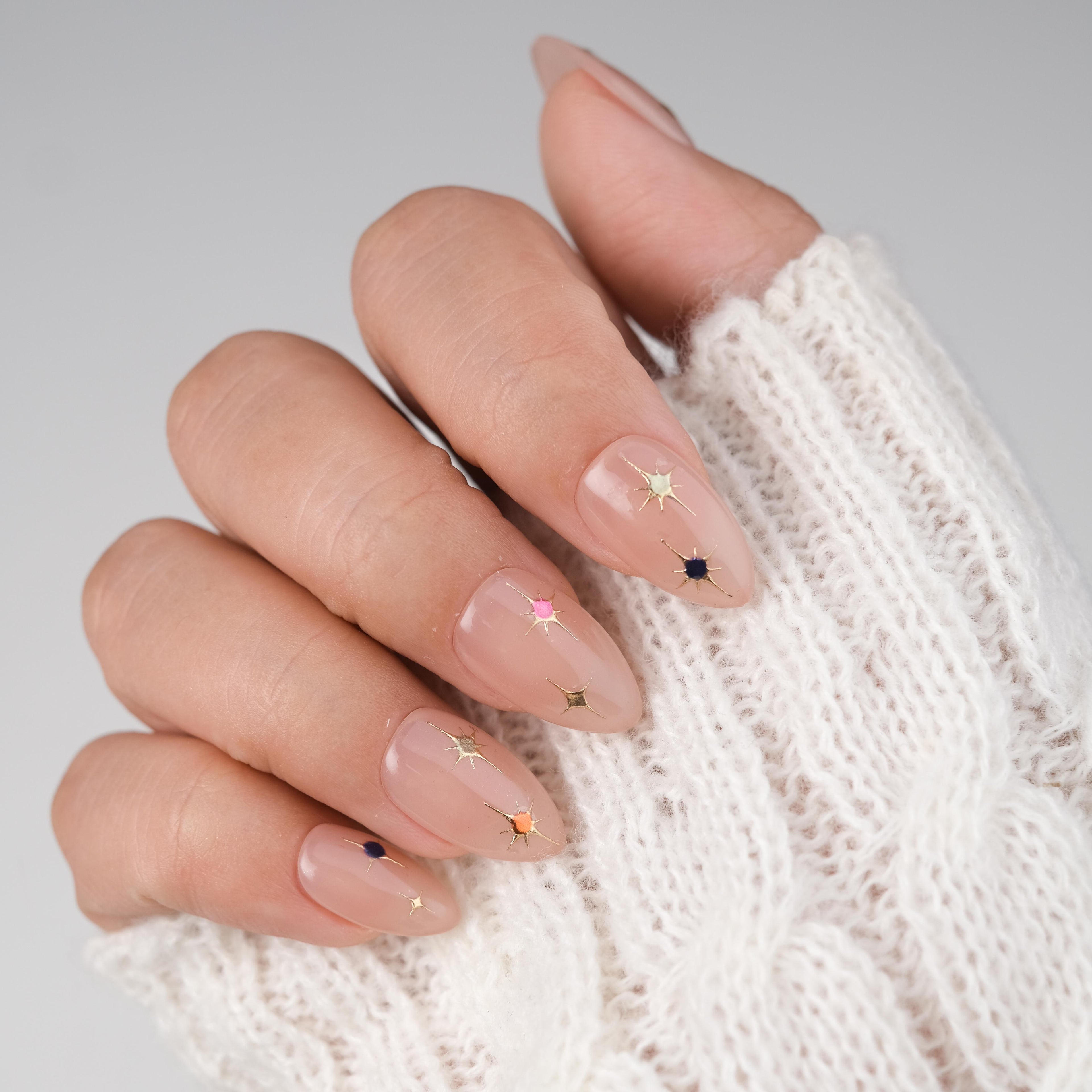Short almond press-on nails by Nailique with nude pink gel finish, gold star accents, and sparkle, reusable with glue and remover.