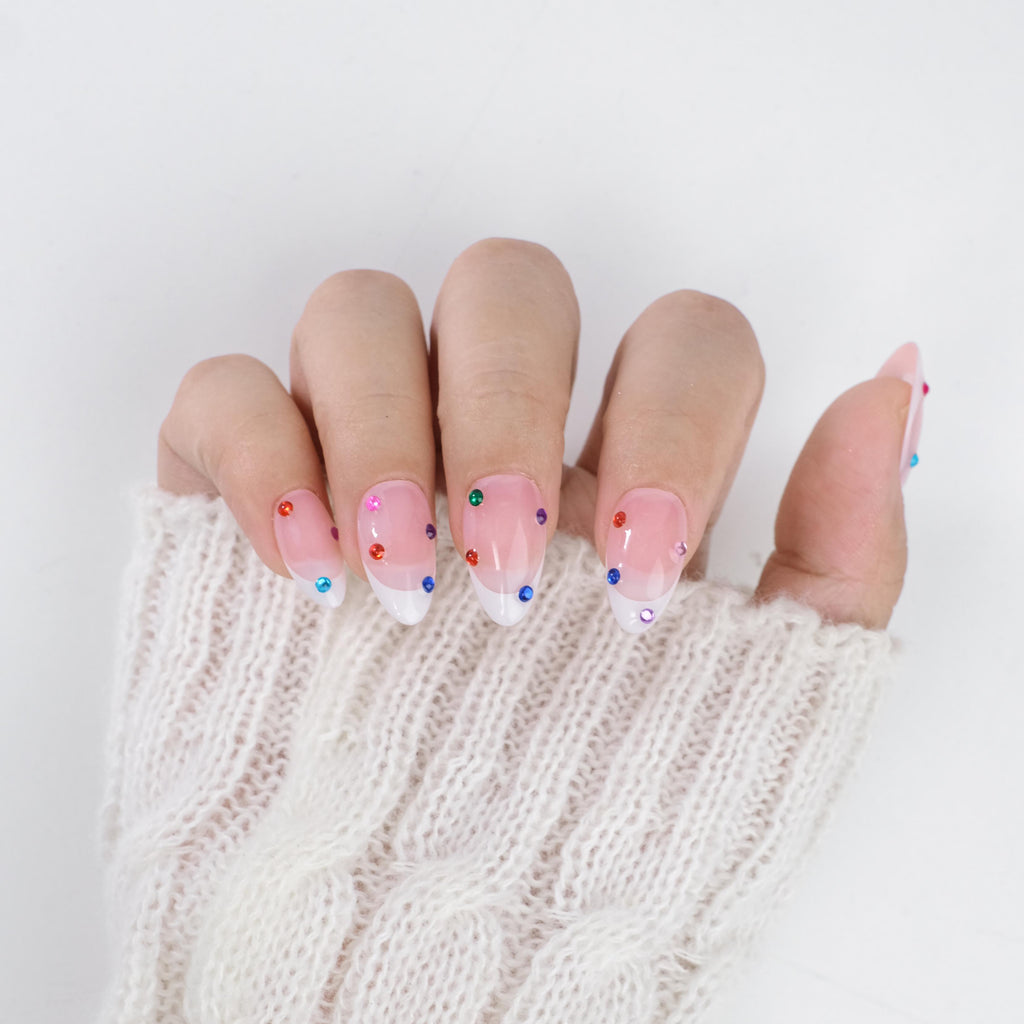 Short almond press-on nails by nailique featuring nude base, white French tips, colorful crystal rhinestones, and a sparkling gel manicure set.